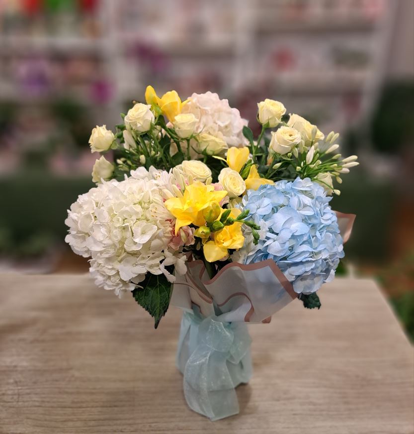 Un buchet alcatuit din hortensii, miniroze si frezii, in vaza de sticla impachetata in hartie impermeabila