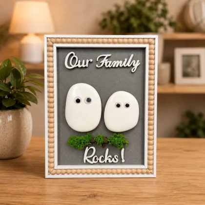 Tablou decorativ handmade „Our Family Rocks” cu pietre decorative, licheni stabilizați și ramă albă cu mărgele din lemn, ideal pentru decorul casei sau cadou.