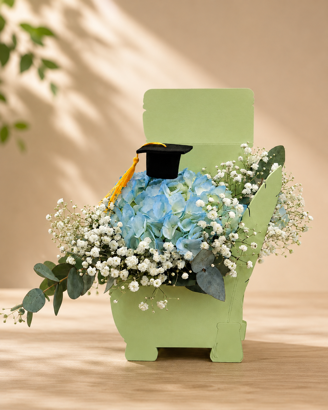 Aranjament floral de absolvire cu hortensie albastră, floarea miresei și tocă decorativă, într-un suport verde elegant.