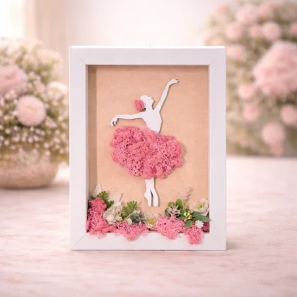 Tablou decorativ cu balerină din licheni stabilizați, în nuanțe roz, cu flori decorative și ramă albă
